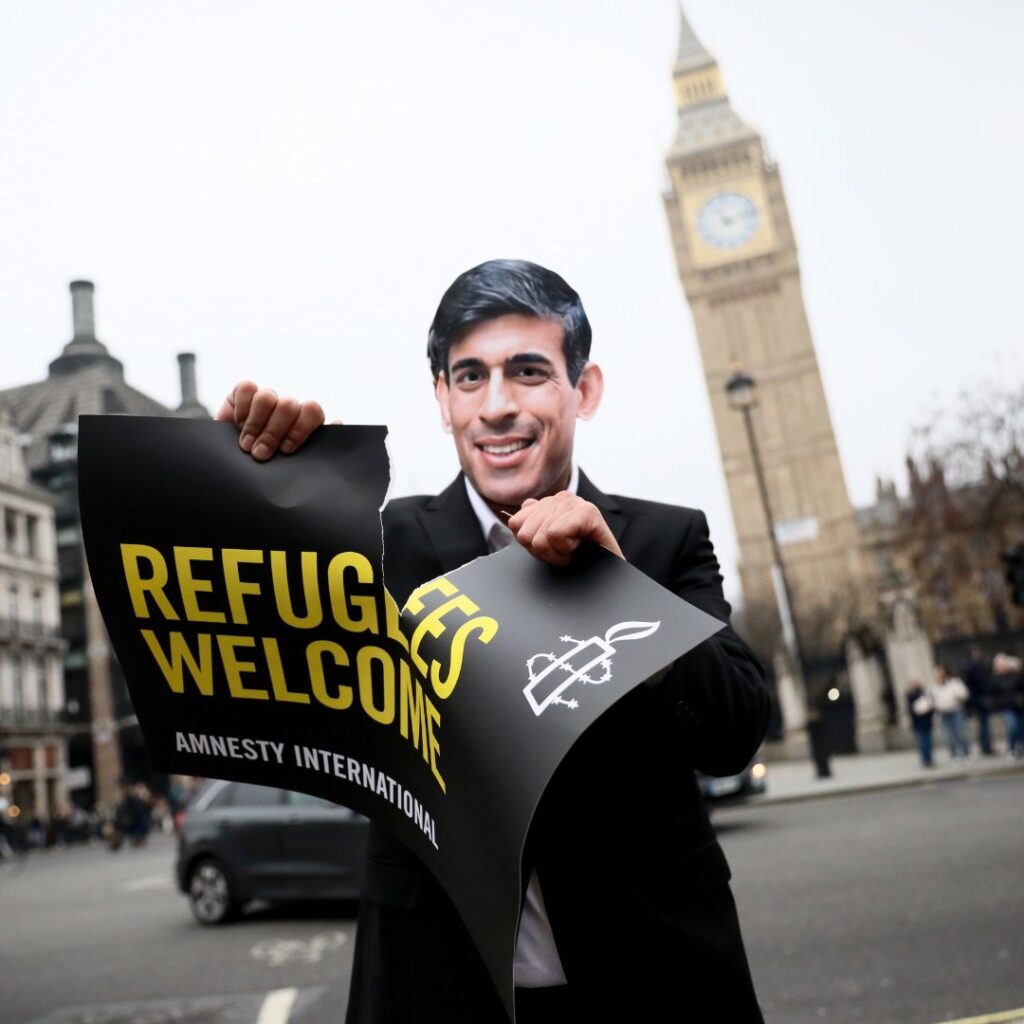 A man in a Rishi Sunak mask rips up a sign that reads Refugees Welcome in front of Big Ben